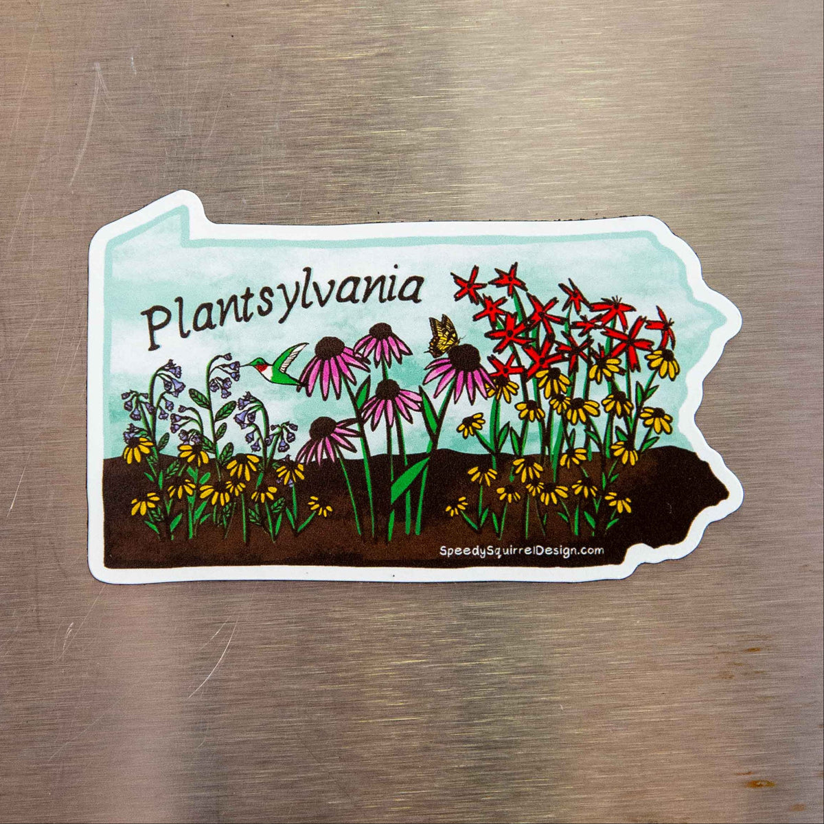 A die-cut sticker shaped like the state of Pennsylvania, featuring a colorful illustration of wildflowers, a hummingbird, and the text 'Plantsylvania' on a brushed metal background.