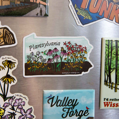 A die-cut sticker shaped like the state of Pennsylvania featuring the text 'Plantsylvania' and an illustration of wildflowers in a field, shown on a stainless steel refrigerator among other magnets.