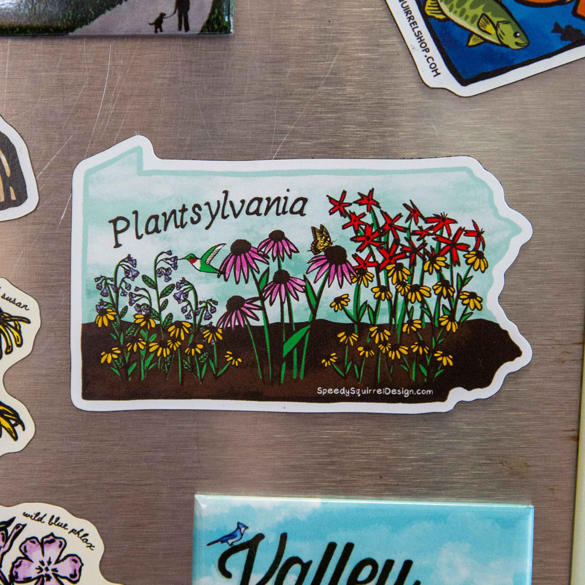 A die-cut sticker shaped like the state of Pennsylvania, featuring a colorful illustration of wildflowers, a hummingbird, and a butterfly with the text 'Plantsylvania' and 'SpeedysSquareDesign.com', shown on a metallic fridge surface.