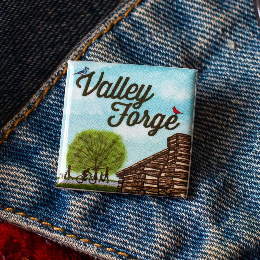Square pin with a Valley Forge Illustration of a log cabin, tree, and people on a bike and footpath with a demim background