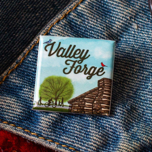 Square pin with a Valley Forge Illustration of a log cabin, tree, and people on a bike and footpath with a demim background