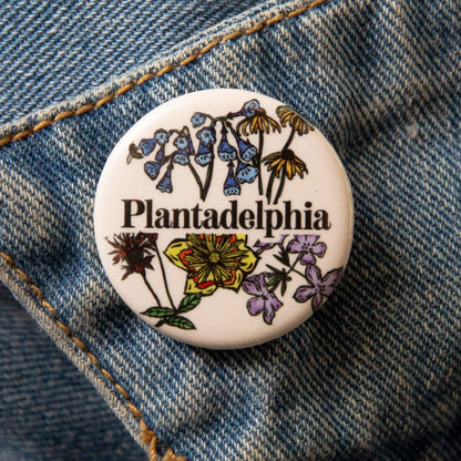 Pin with 'Plantadelphia' text and floral designs on a denim background