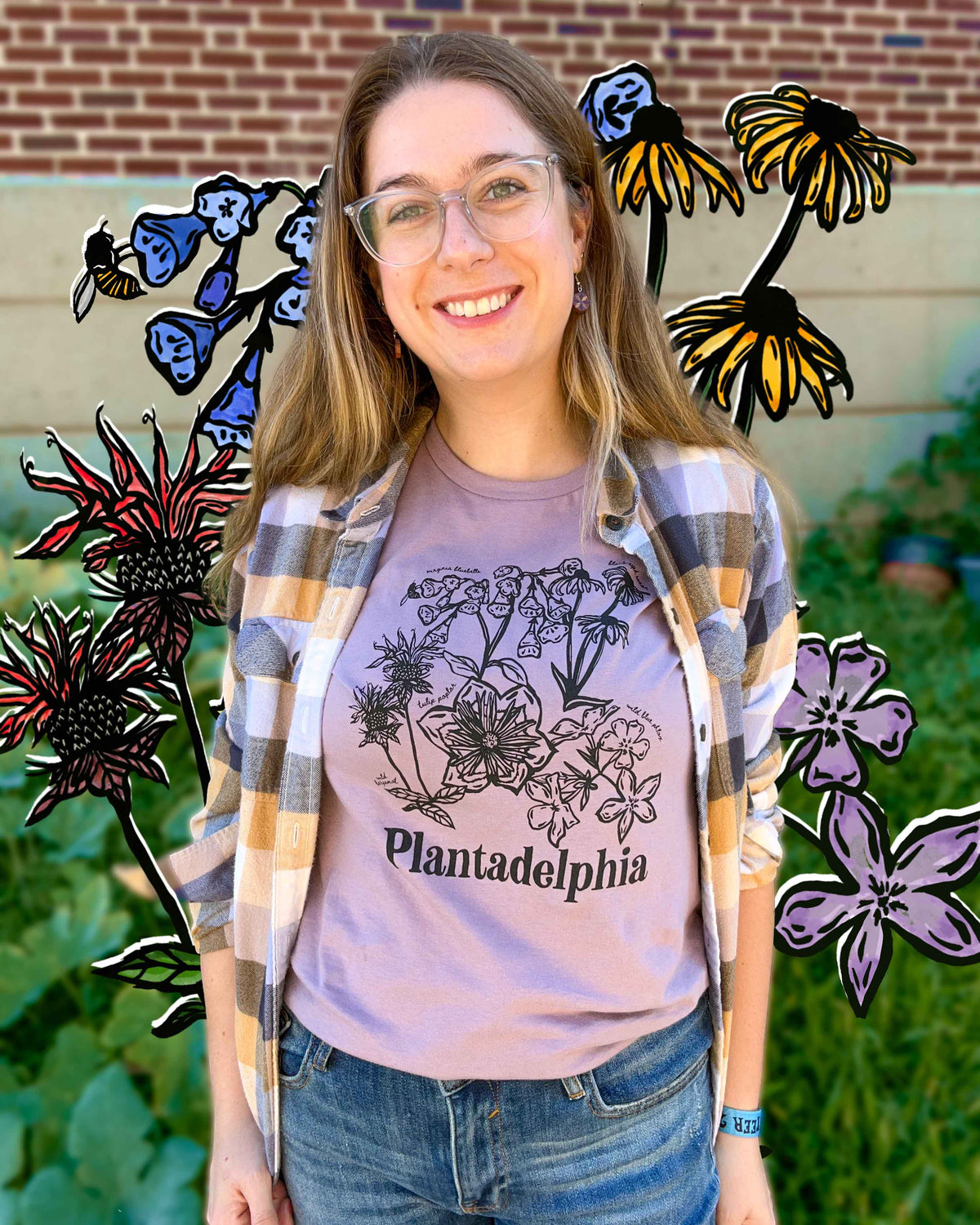A young woman smiling wearing a light mauve-pink t-shirt with a black botanical illustration and the text 'Plantadelphia' printed on the front, photographed outdoors against a brick wall and greenery with the colorful floral illustration behind her. 