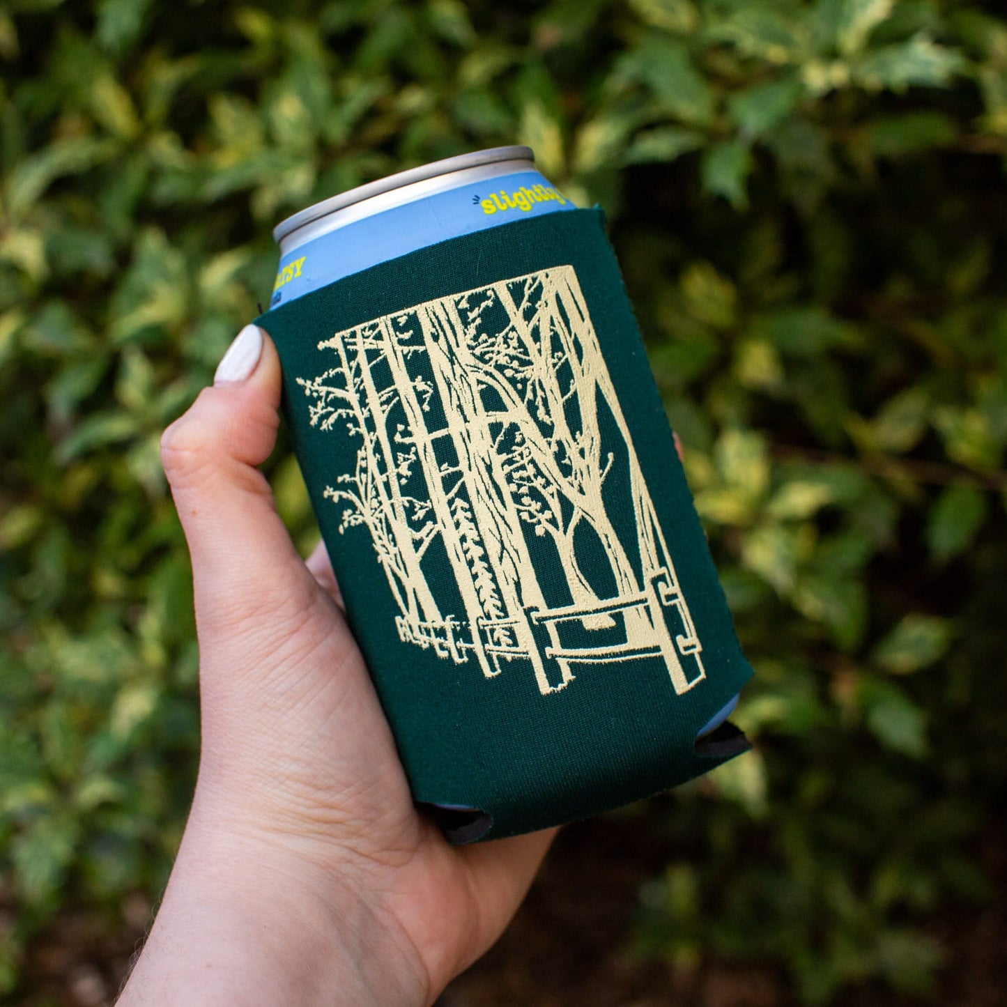 Hand holding a can with a green sleeve featuring a cream design against a blurred green background