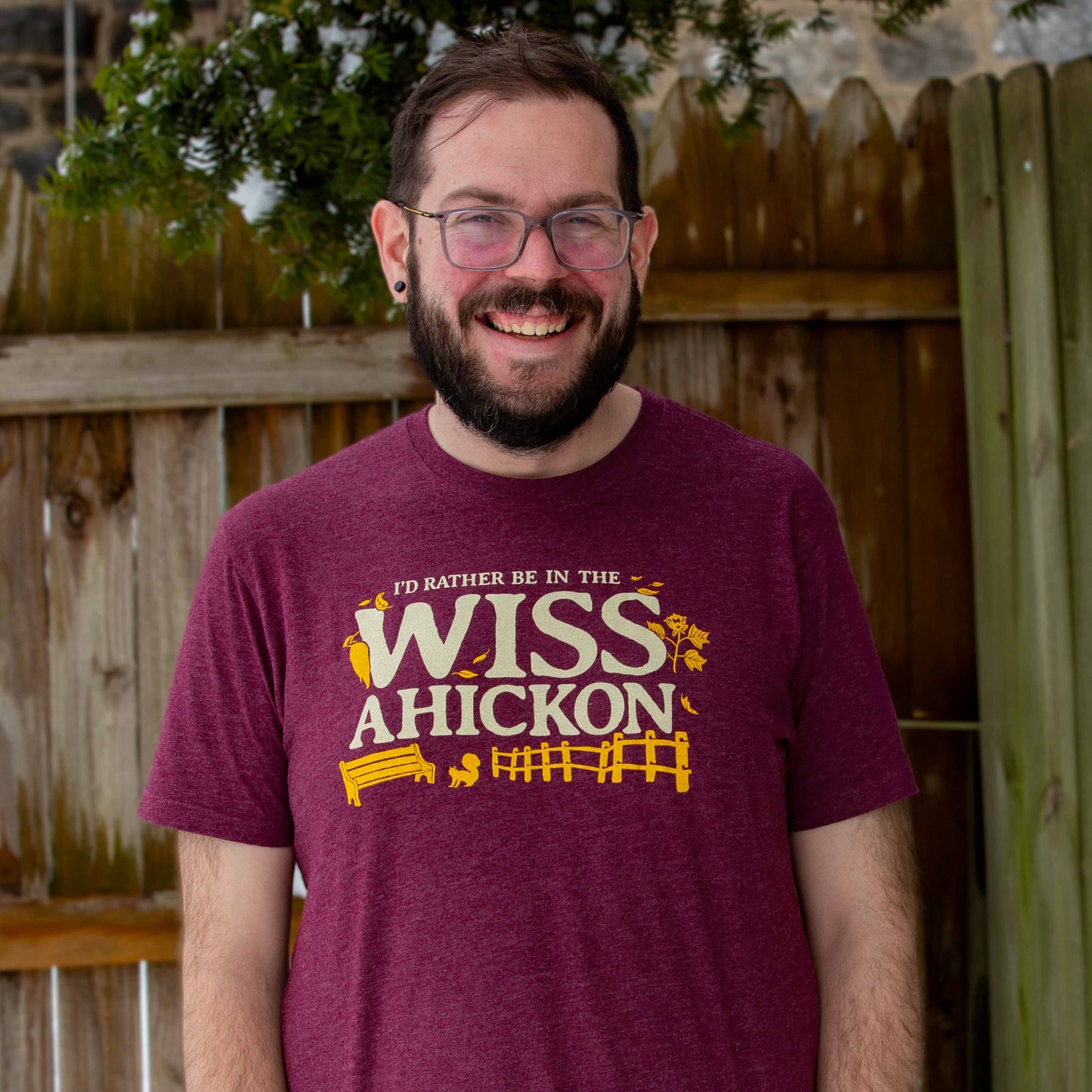 Man wearing a maroon t-shirt with 'I'd rather be in the Wissahickon' text and graphics outdoors.