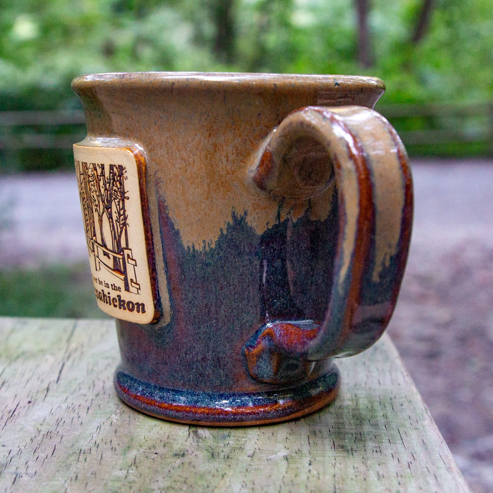 Handmade ceramic mug with a handle and a small sign on a wooden table, outdoors.