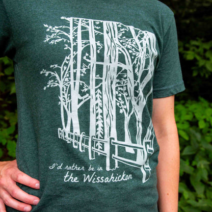 Young woman wearing a green t-shirt with a light green fence and forest with text 'I'd rther be in the Wissahickon' while standing outdoors.