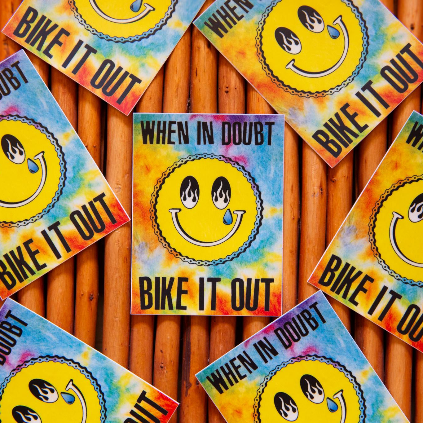 Multiple stickers with a tie-dye pattern and a smiling face design that includes a flame and a tear drop. Each sticker has the text 'BIKE IT OUT' on it.