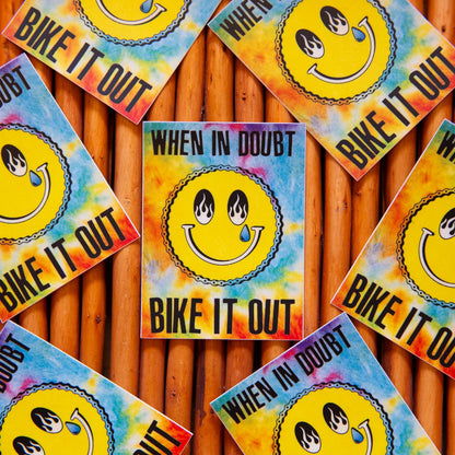 Multiple stickers with a tie-dye pattern and a smiling face design that includes a flame and a tear drop. Each sticker has the text 'BIKE IT OUT' on it.